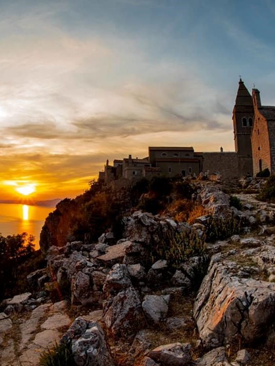 Sonnenuntergang hinter einer Festung auf einer Klippe, umgeben von groben Felsen.